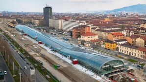 Une gare moderne dotée d’un toit incurvé en verre, entourée de bâtiments urbains et avec une chaîne de montagnes en toile de fond.