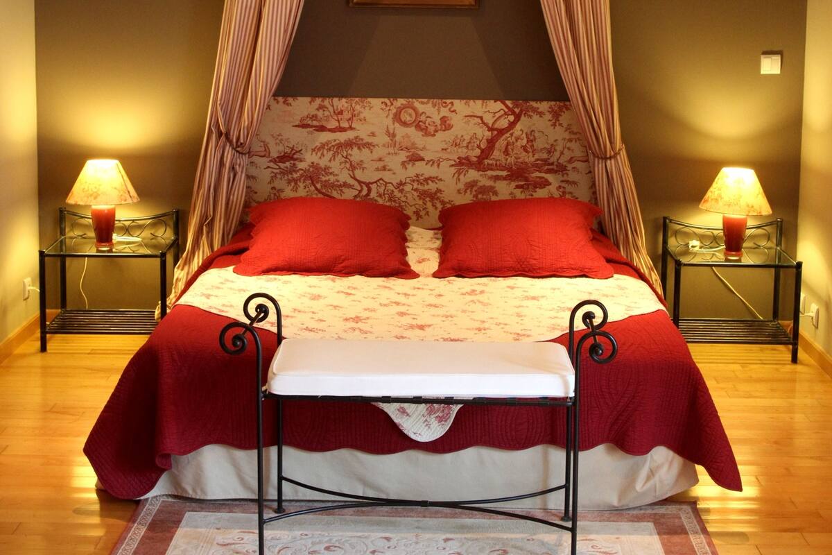 Un dormitorio con cama de dosel, dos mesitas de noche con lámparas, una colcha roja y un tapete decorativo.