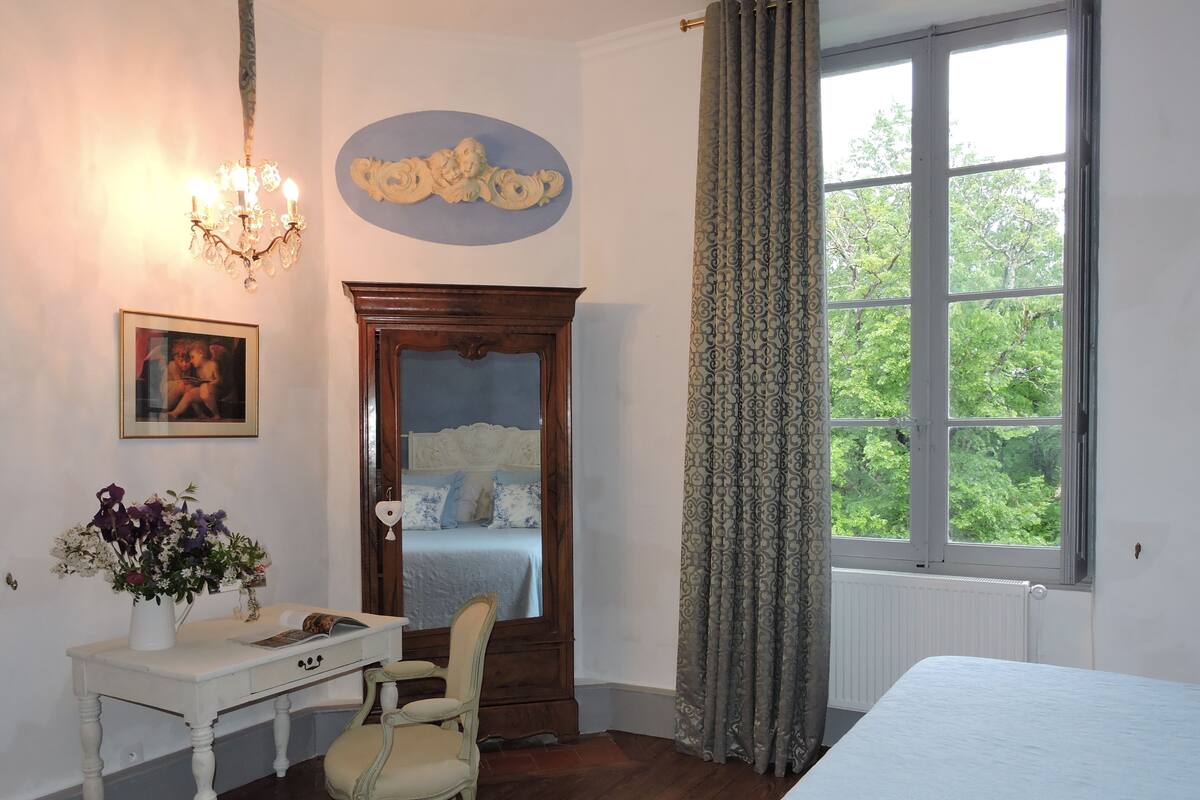 Un dormitorio con cama de madera, mesita de noche, silla, cómoda, lámpara de araña, ventana con cortinas y decoración en la pared.