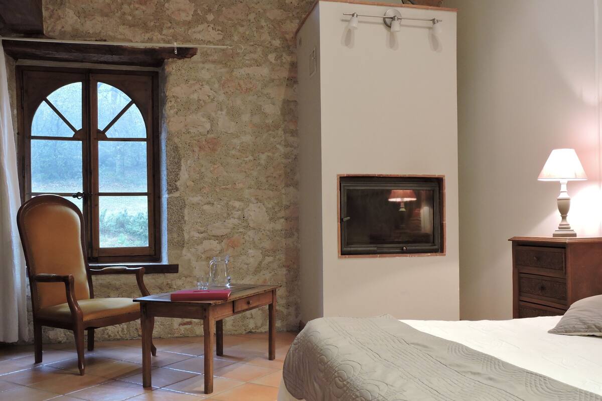 Un dormitorio con cama, una silla, una mesa, una lámpara y una ventana que ofrece vistas a árboles.