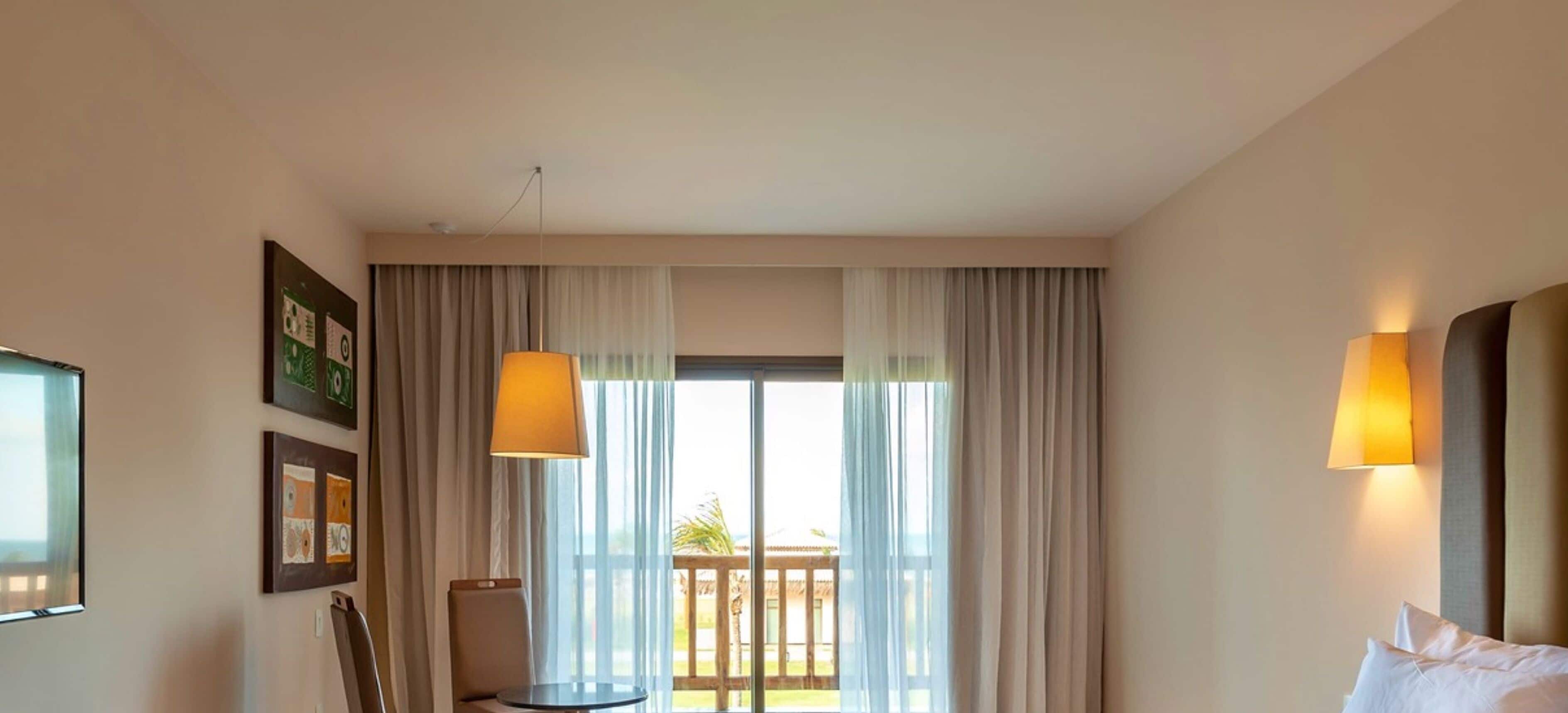 A hotel room with a bed, a chair, a table, and a balcony with curtains.