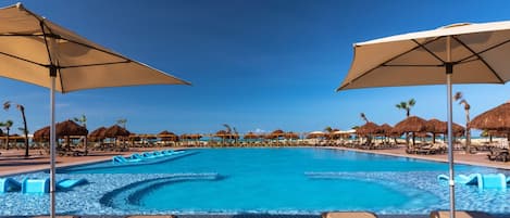 Una amplia piscina al aire libre con sillones de playa y sombrillas, rodeada de palmeras y construcciones con techo de paja.