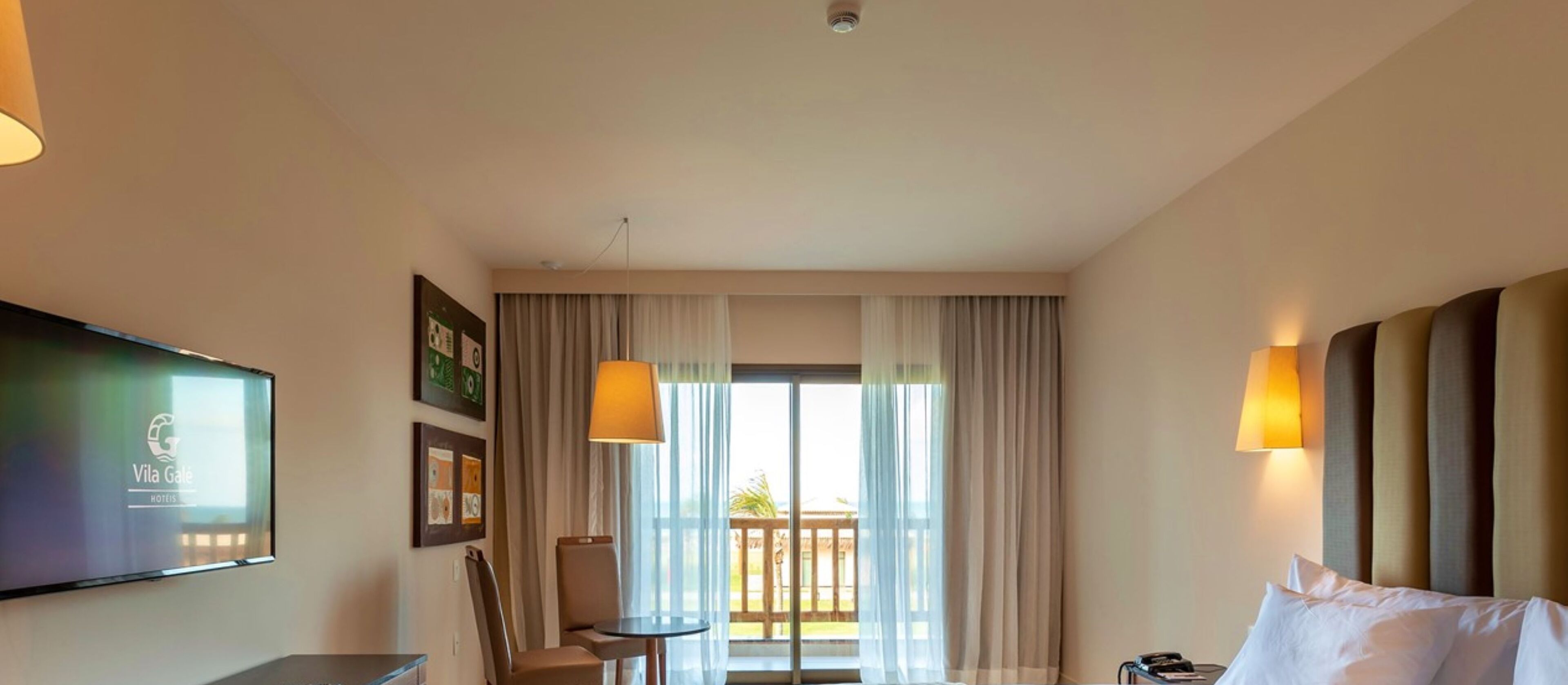 A hotel room with a bed, a TV mounted on the wall, a small table, and a chair. There is a window with curtains and a view of the outside.