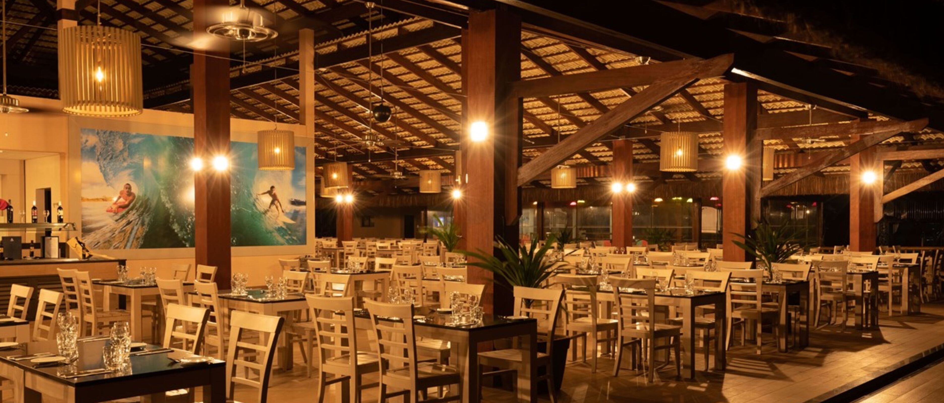 A spacious restaurant with high ceilings and wooden beams, featuring multiple dining tables and chairs, and a large mural on the wall.