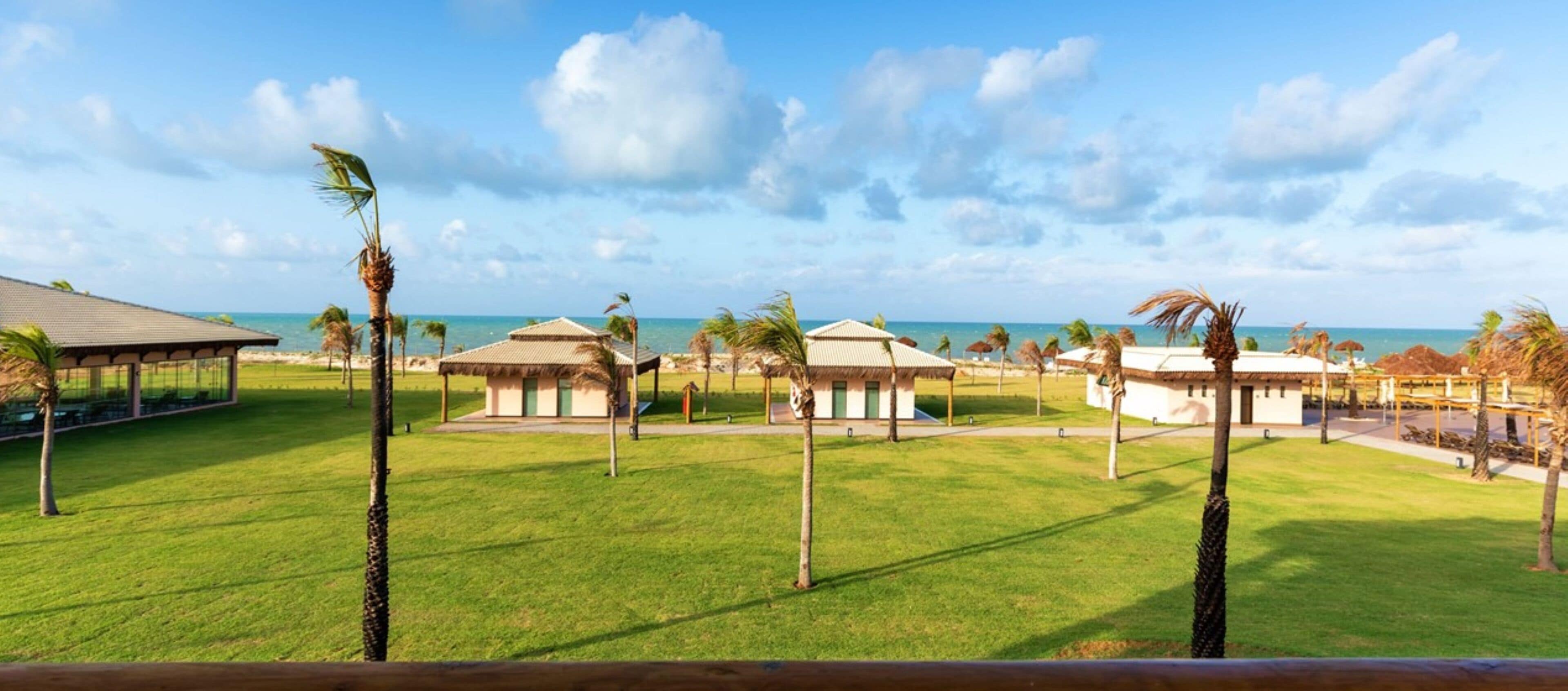 Un resort con varias construcciones de techo de paja, un césped y una vista despejada del océano.