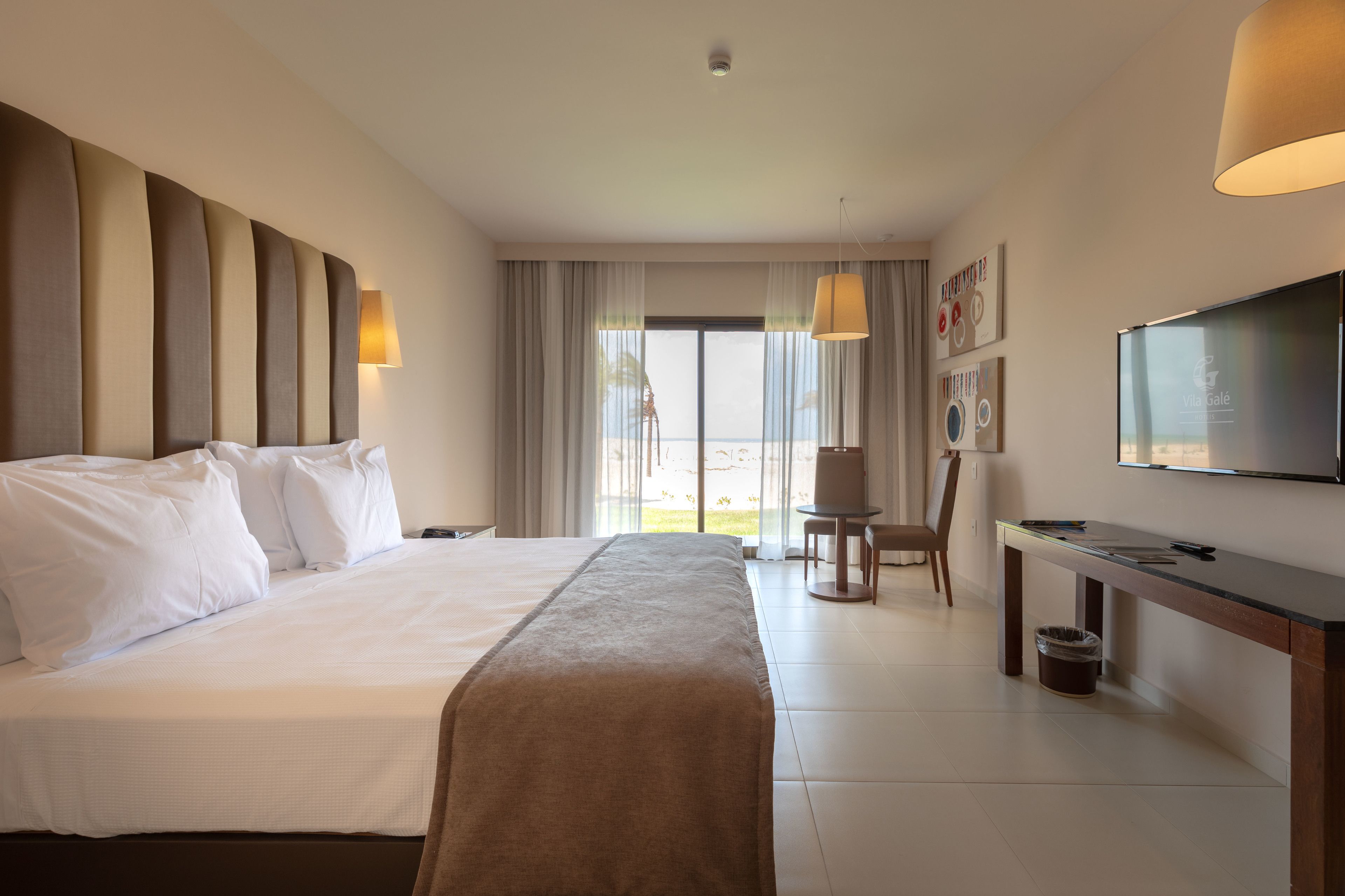 A hotel room with a large bed, a desk, a chair, a TV, and a view of the outdoors.