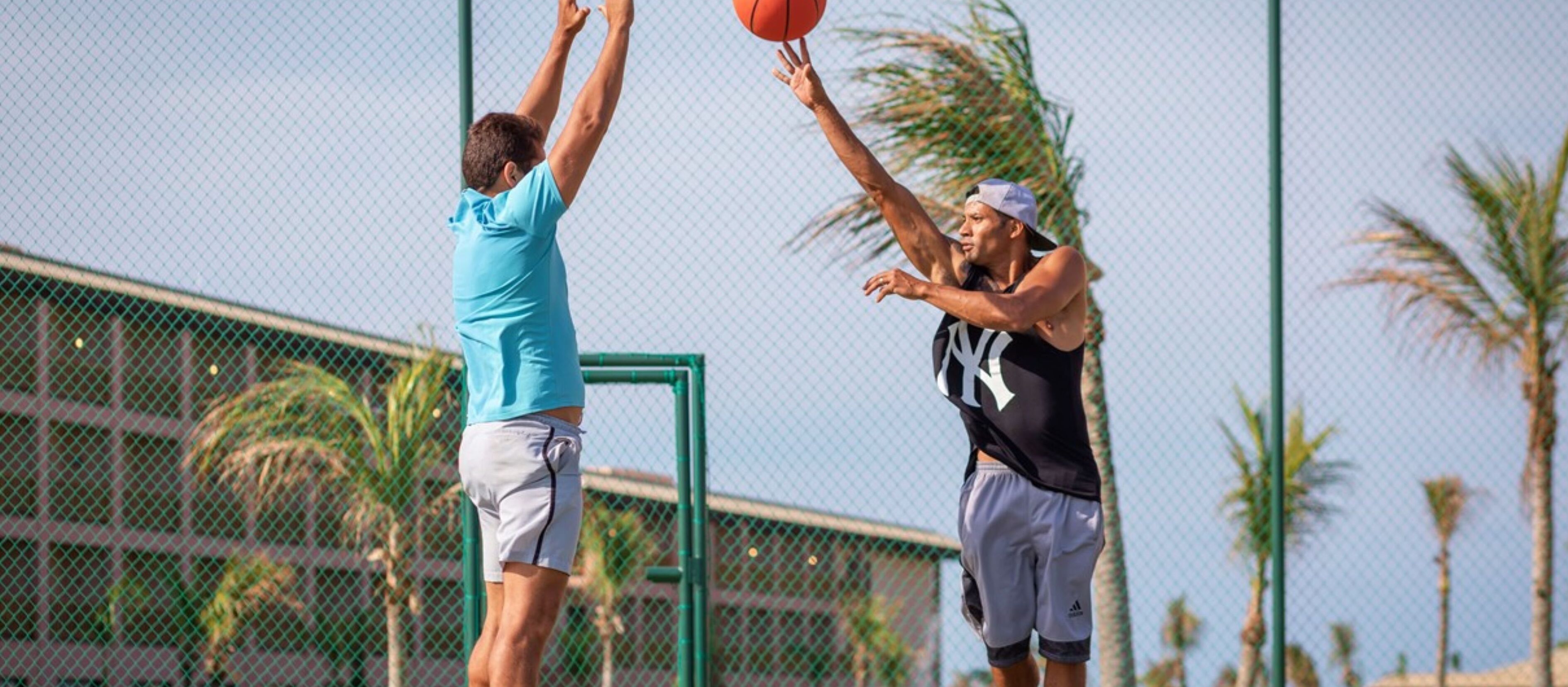 Two people playing basketball on an outdoor court with a chain-link fence and palm trees in the background.
