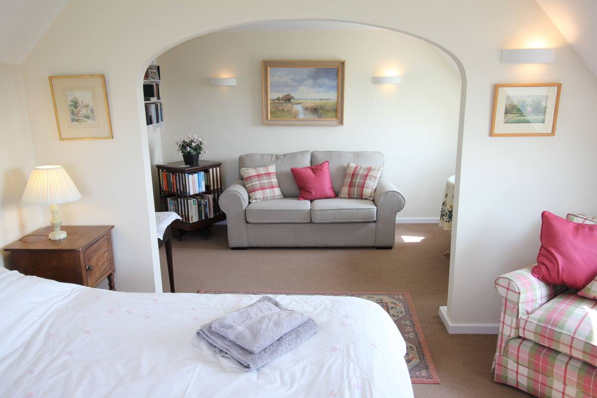 A bedroom with a bed, a grey sofa, a small armchair, a bookshelf, and a painting on the wall.