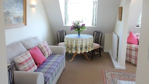 Una habitación acogedora con un sofá blanco, una mesita redonda con mantel floreado y una ventana con cortinas.