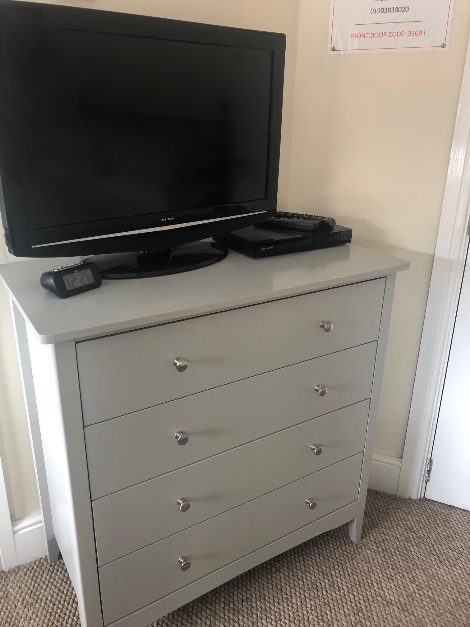 A grey dresser with a flat-screen TV and a DVD player on top, a wall-mounted sign with a door code, and a carpeted floor.