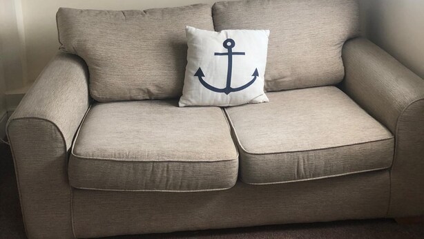 Un canapé beige agrémenté d’un coussin orné d’un motif d’ancre, placé dans une pièce dont le plafond est en pente et où l’on peut voir un tableau accroché au mur.
