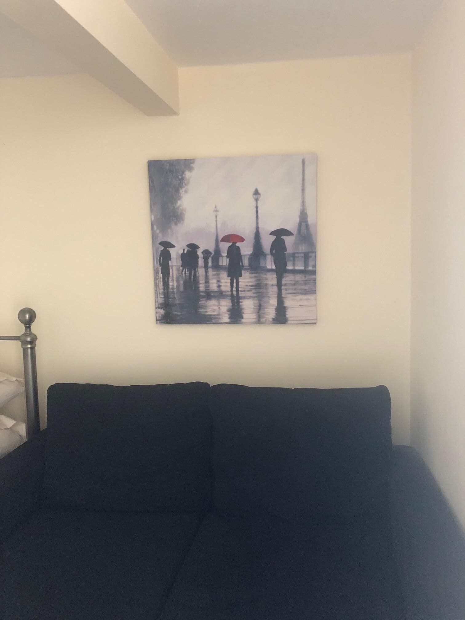 A room with a dark-colored sofa against a wall, a painting of a rainy Paris street scene, and a metal bed frame.