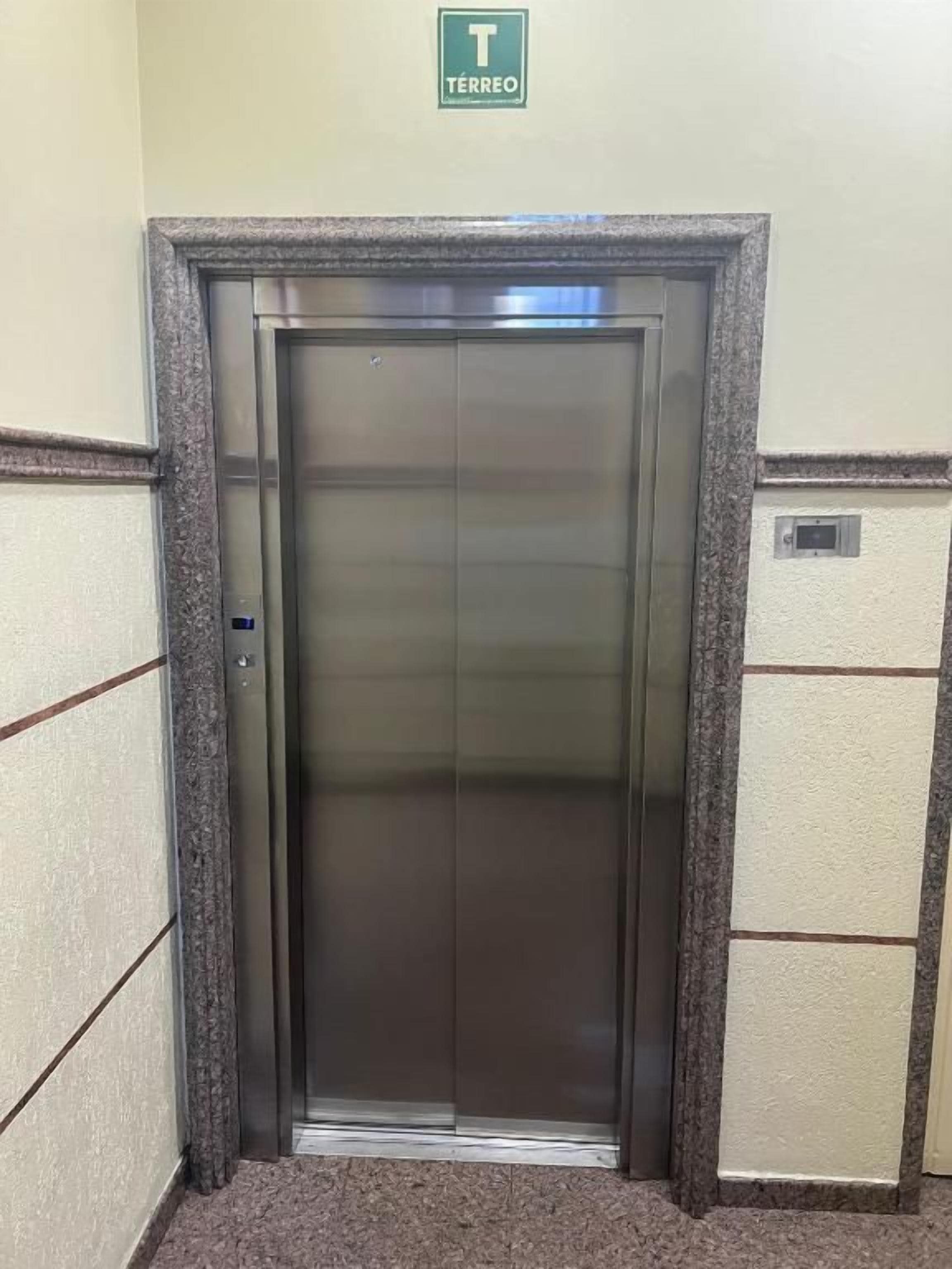 Een lift met een metalen afwerking, een groen bord erboven en een gestructureerde wand aan weerszijden.