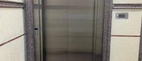 Een lift met een metalen afwerking, een groen bord erboven en een gestructureerde wand aan weerszijden.