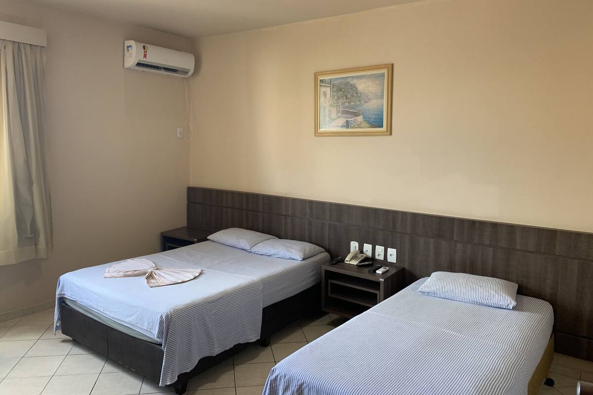 A hotel room with two single beds, a nightstand with a phone and remote controls, an air conditioning unit, and a painting on the wall.