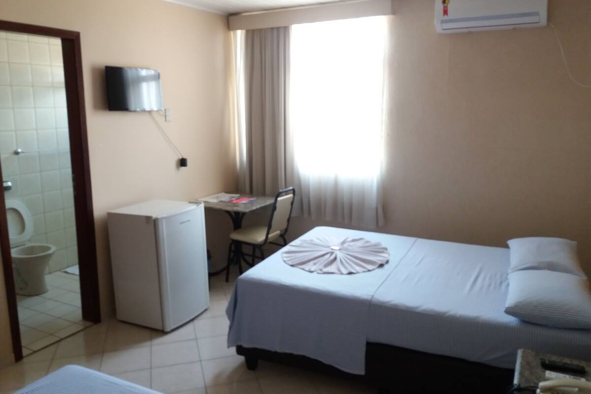 A hotel room with a bed, a desk, a chair, a small refrigerator, a television, and an air conditioning unit.