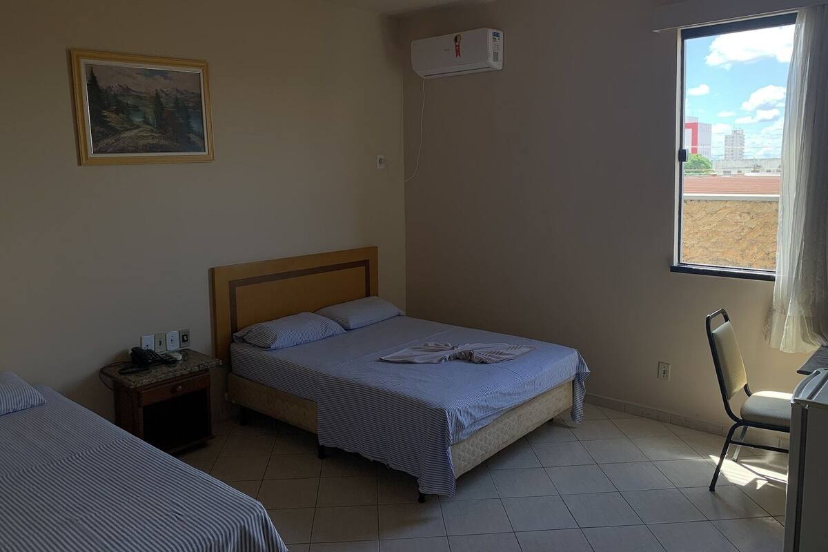 A hotel room with a bed, a nightstand, a chair, a window with a view, and an air conditioning unit.