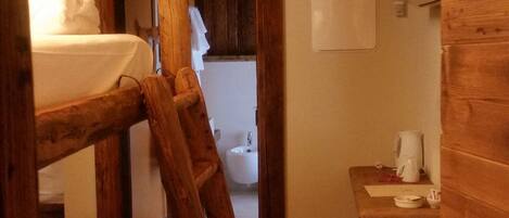 A rustic wooden room with a bunk bed, a ladder, a small stool, and a desk with a lamp.
