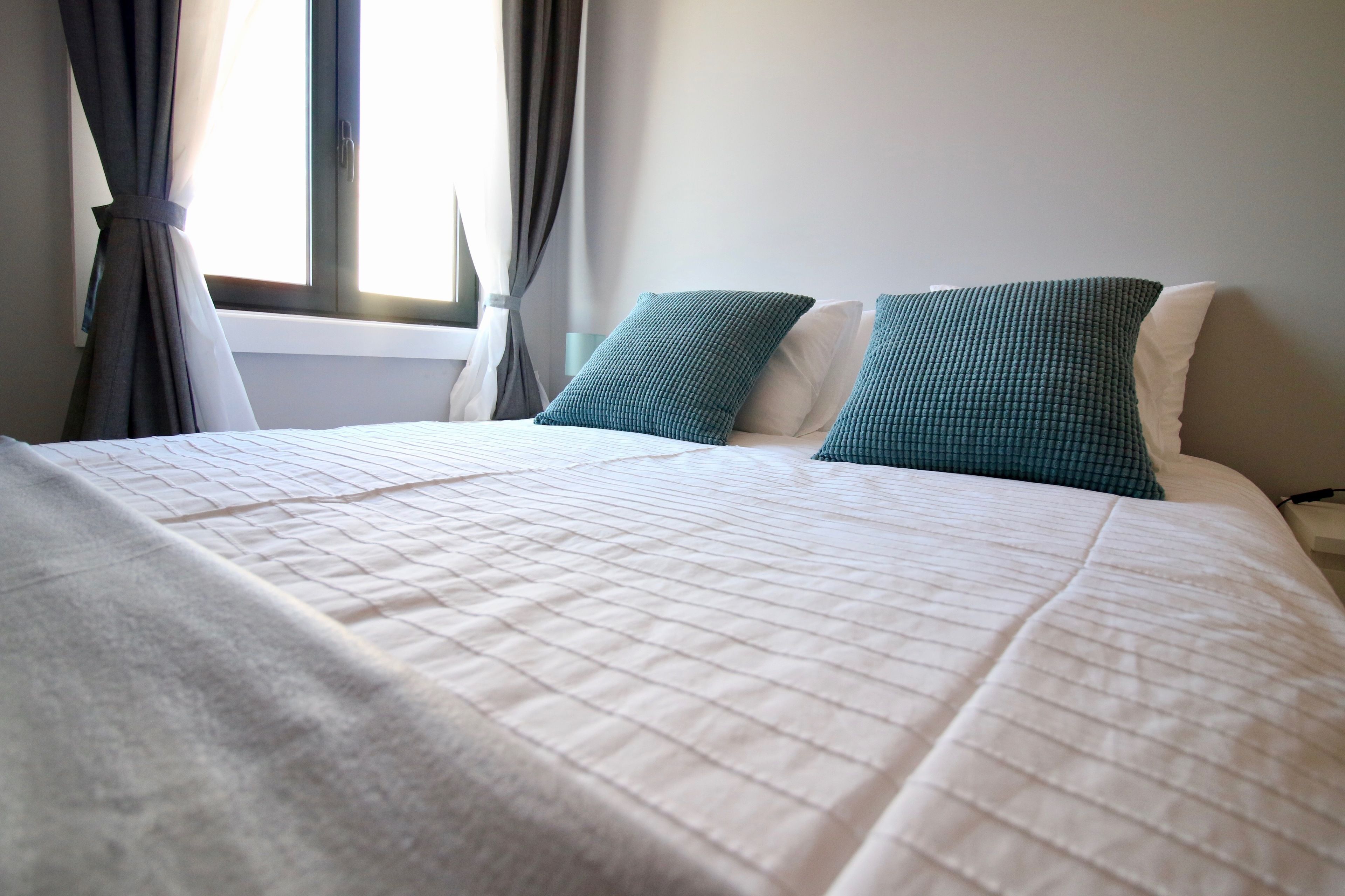 Ein ordentlich bezogenes Bett mit weißer gesteppter Tagesdecke und zwei Kissen, eines türkis und eines weiß, in einem Zimmer mit Fenster und Vorhängen.