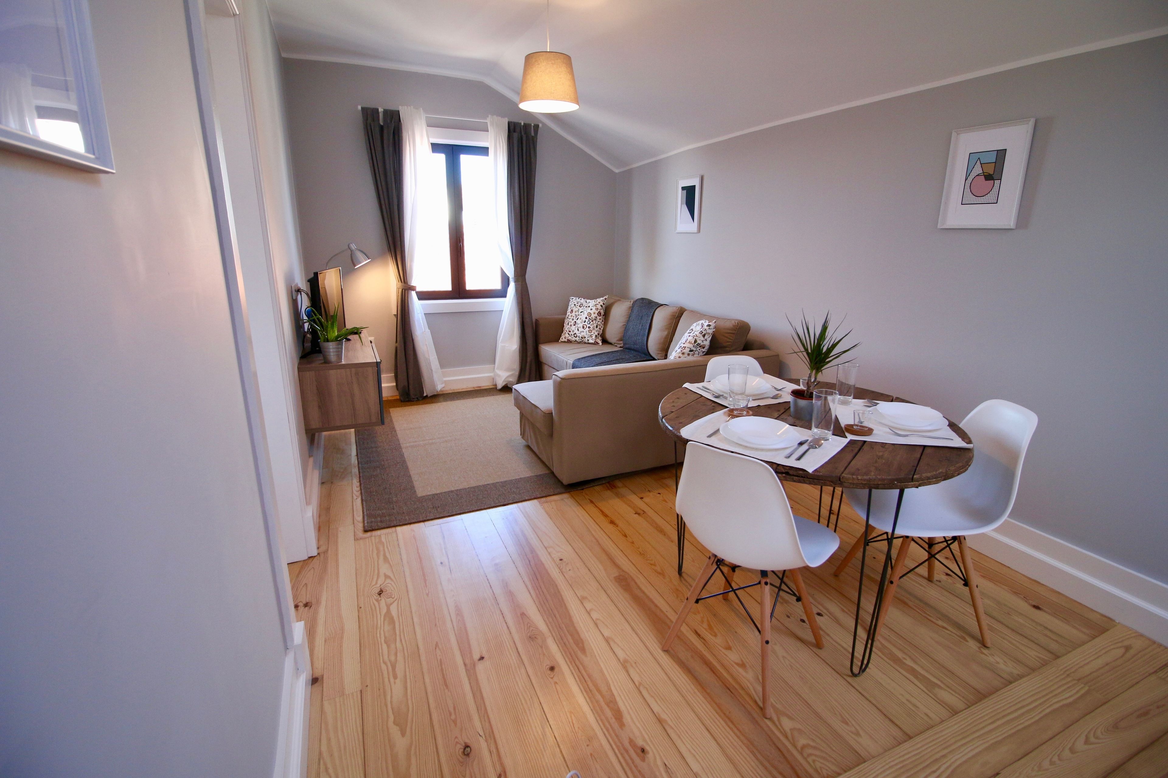 A modern living room with a wooden floor, a sofa, a dining table with chairs, and a small side table with a lamp.