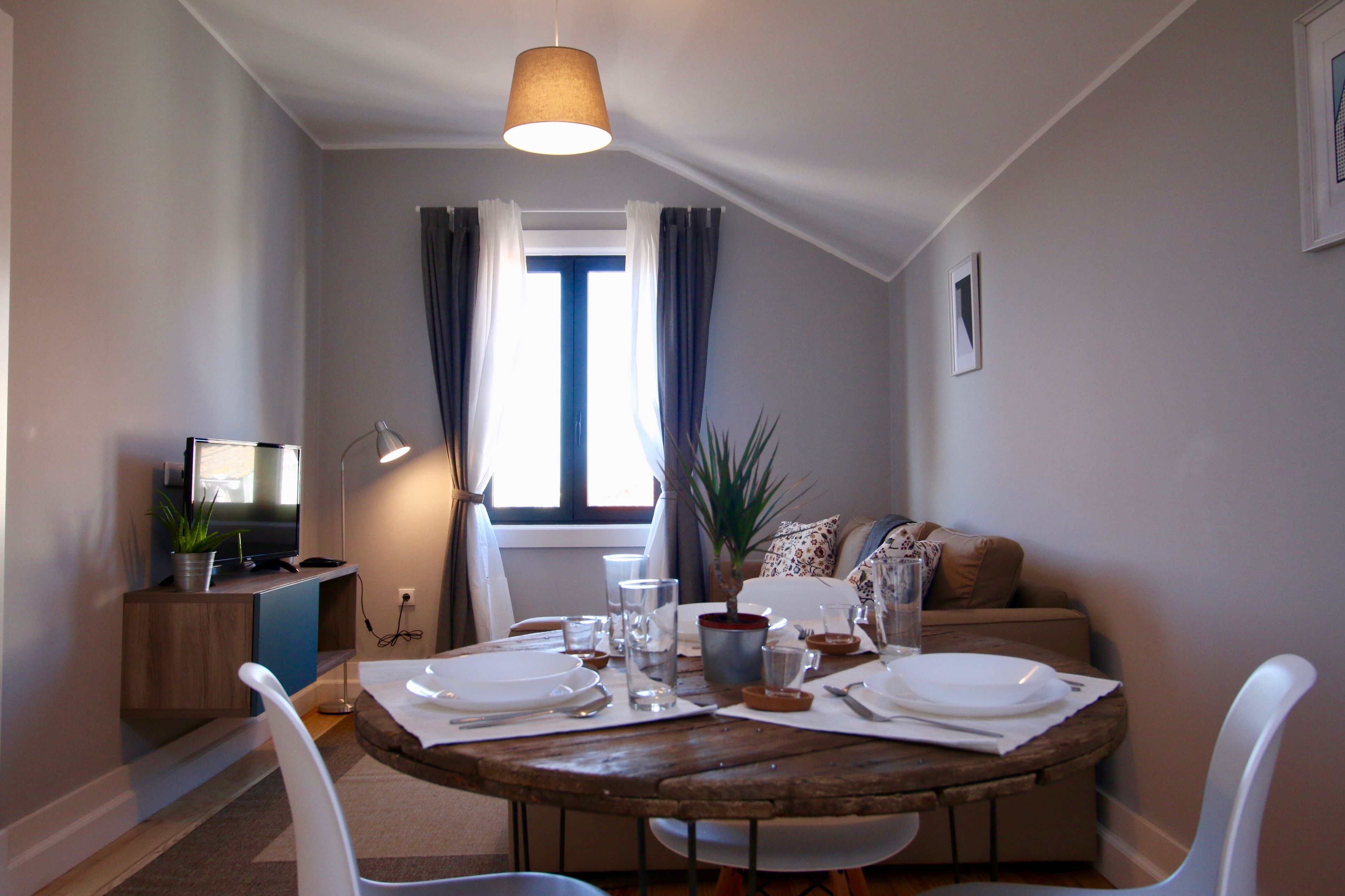 Un comedor con una mesa redonda de madera preparada para dos personas, un televisor sobre un mueble bajo, una planta y una ventana con cortinas.
