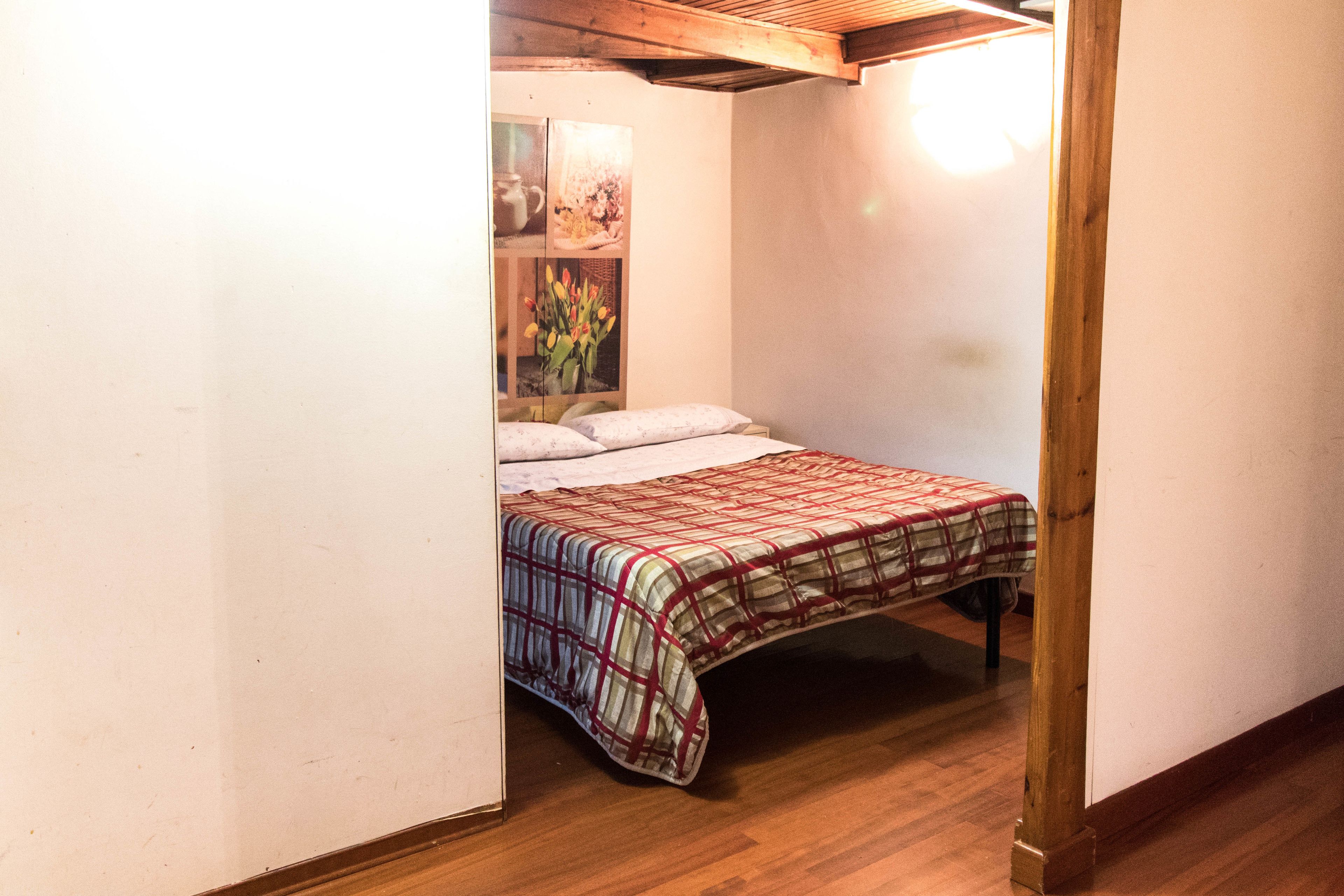 A bedroom with a wooden floor, a bed with a plaid bedspread, and a wall with a picture of flowers.