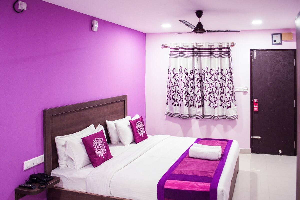 A hotel room with a purple wall, a bed with white and purple bedding, a ceiling fan, and a door with a lock.