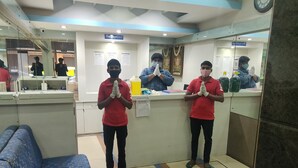 A reception area with two individuals standing in prayer, a reception desk, a blue patterned couch, and a tiled floor with color stripes. - OYO 8501 Abhimaani Comforts (Bengaluru)
