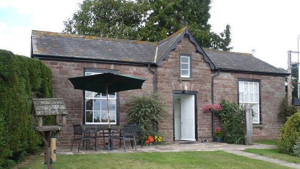 A brick house with a garden, a wooden bird feeder, and a table with chairs under a green umbrella. - Garden Cottage & Granary (Ross-on-Wye)