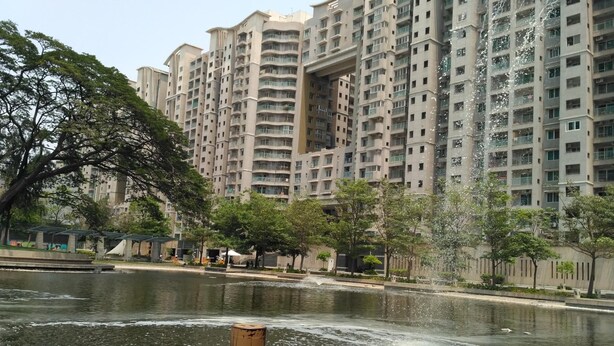 A high-rise residential building with multiple balconies, a water feature in the foreground, and a park area with trees and a small pond. - OYO 9253 Champion Comfort (Bengaluru)