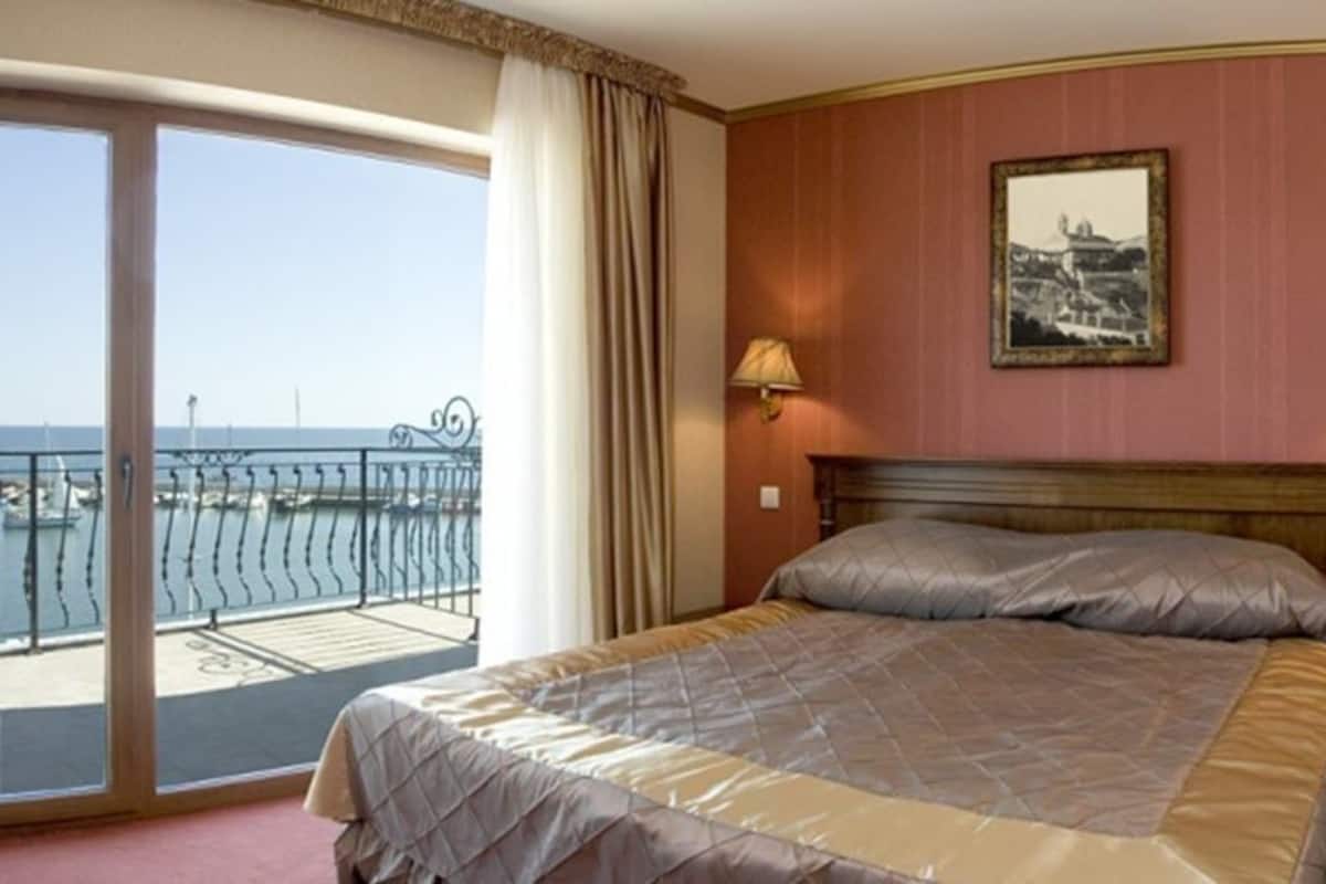 A hotel room with a bed, a balcony with a view of the sea, and two wall-mounted lamps.