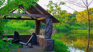 A gazebo with a stone base and a wooden roof, situated by a calm pond surrounded by lush greenery.