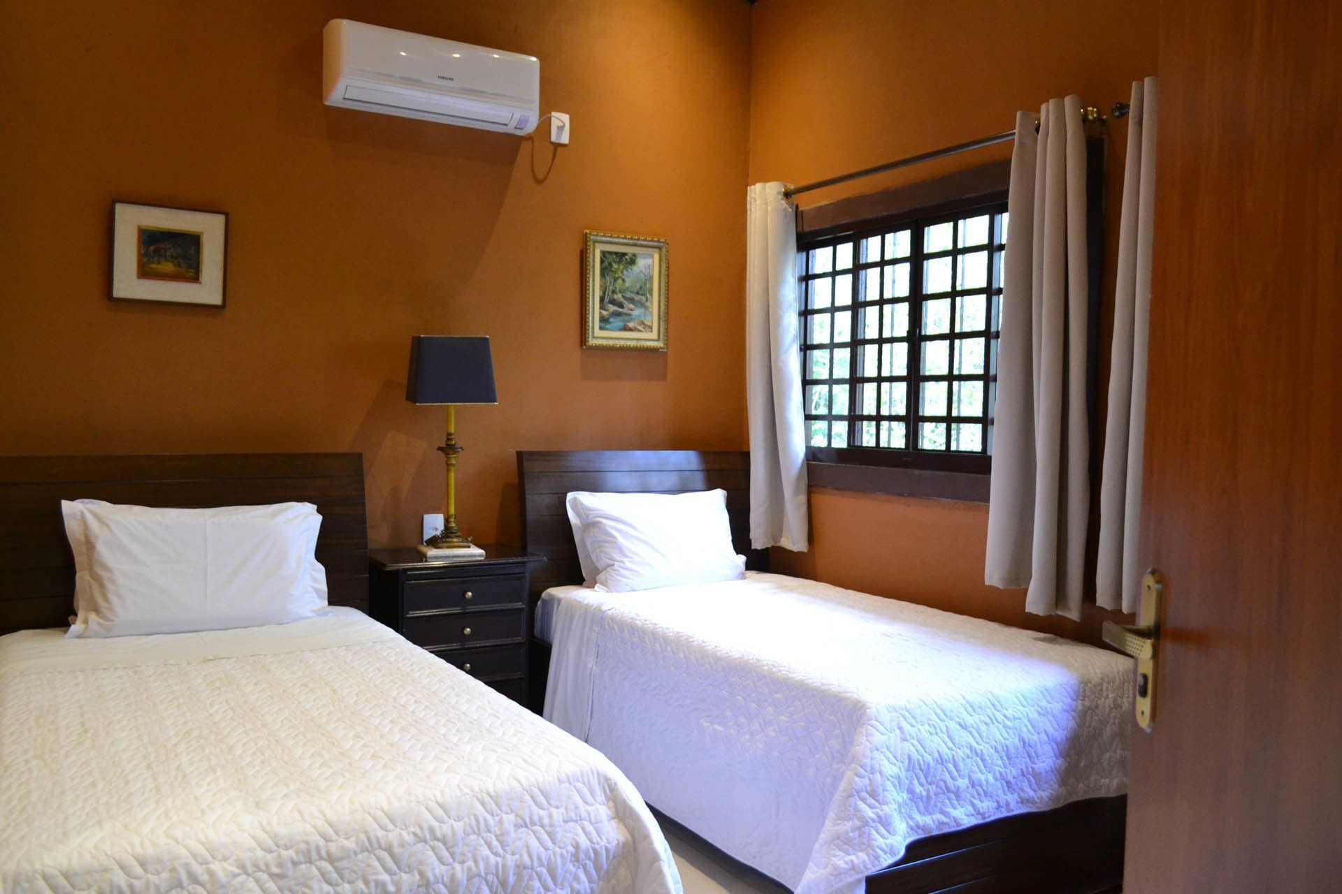Une chambre d’hôtel avec deux lits, une table de chevet, une lampe, un climatiseur et une fenêtre avec des rideaux.