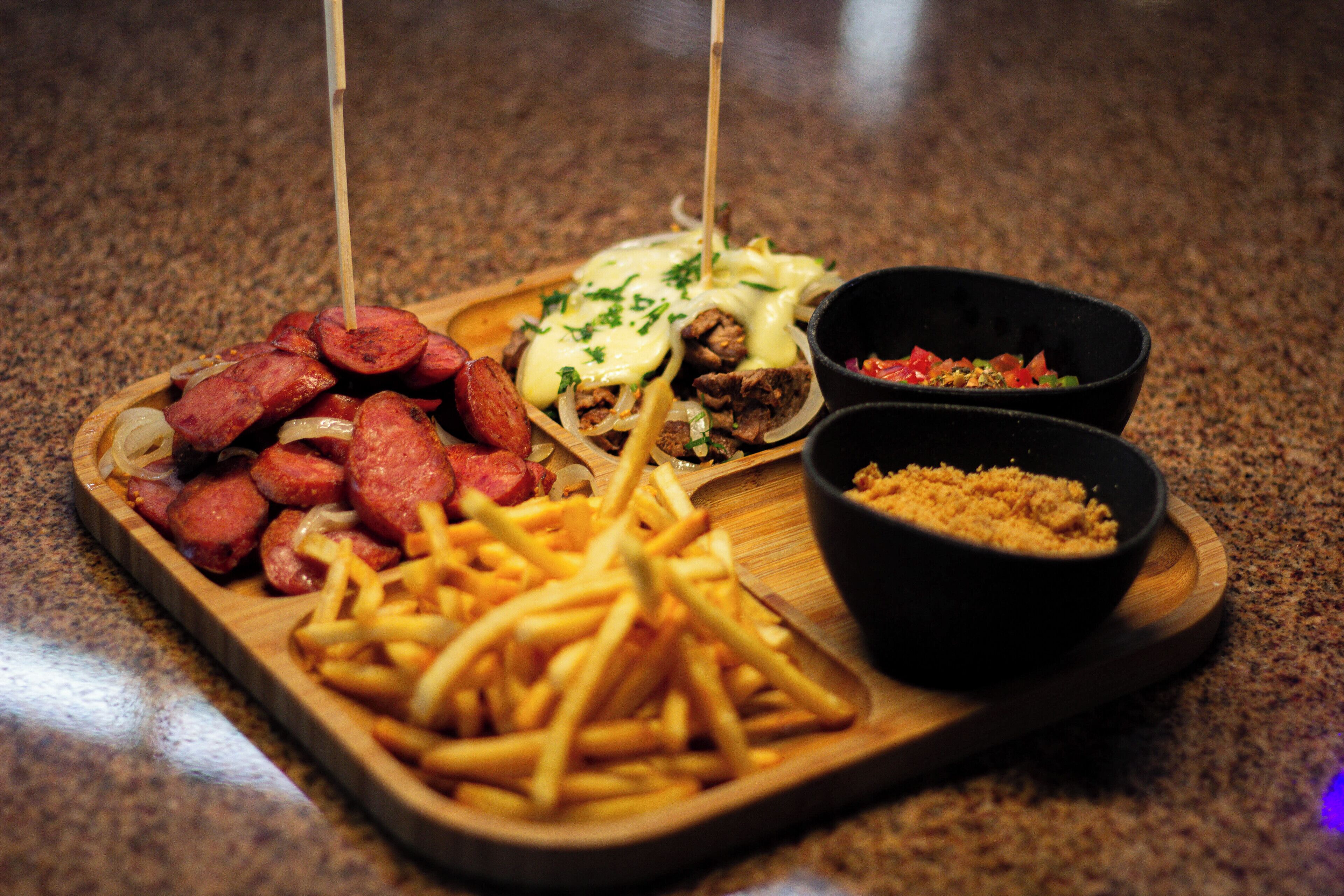 A wooden tray with various foods including sausages, fries, and two bowls of sauces.
