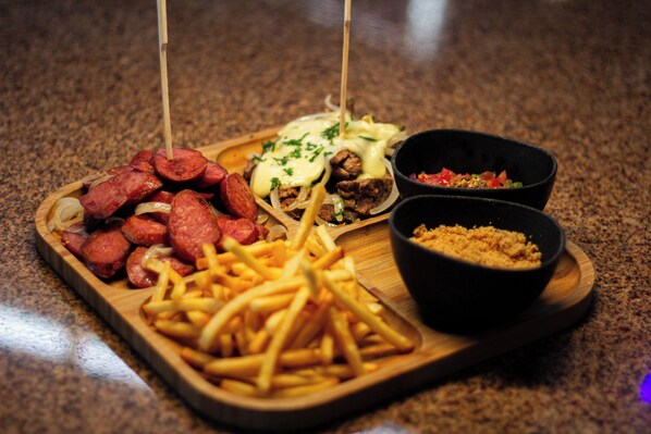 A wooden tray with various foods including sausages, fries, and two bowls of sauces.