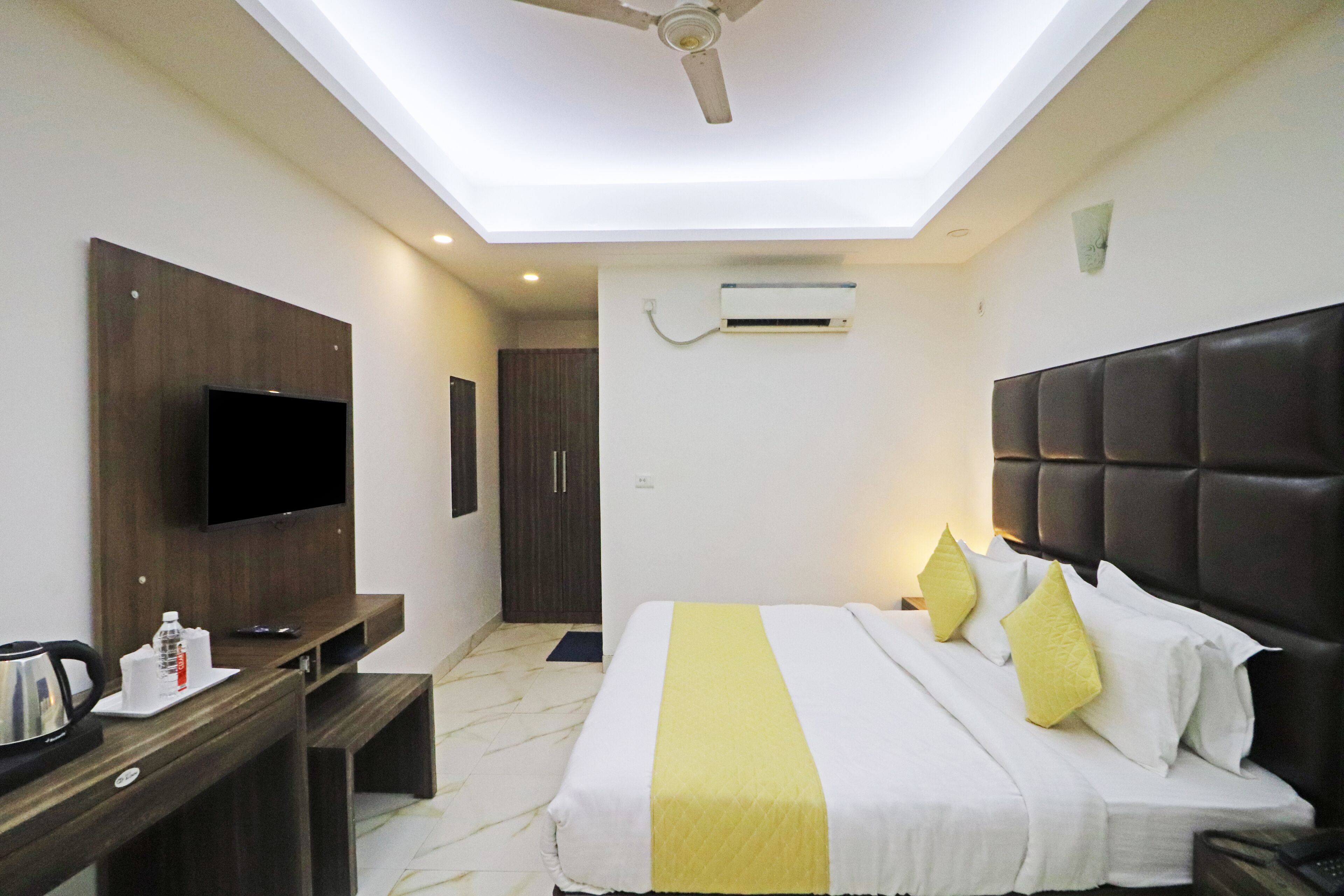 A hotel room with a bed, a TV mounted on the wall, a ceiling fan, an air conditioning unit, and a desk with a kettle and a bottle.