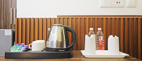 Habitación de hotel con pared de paneles de madera, cafetera, taza, bandeja con una botella y pañuelos, y un refrigerador pequeño.