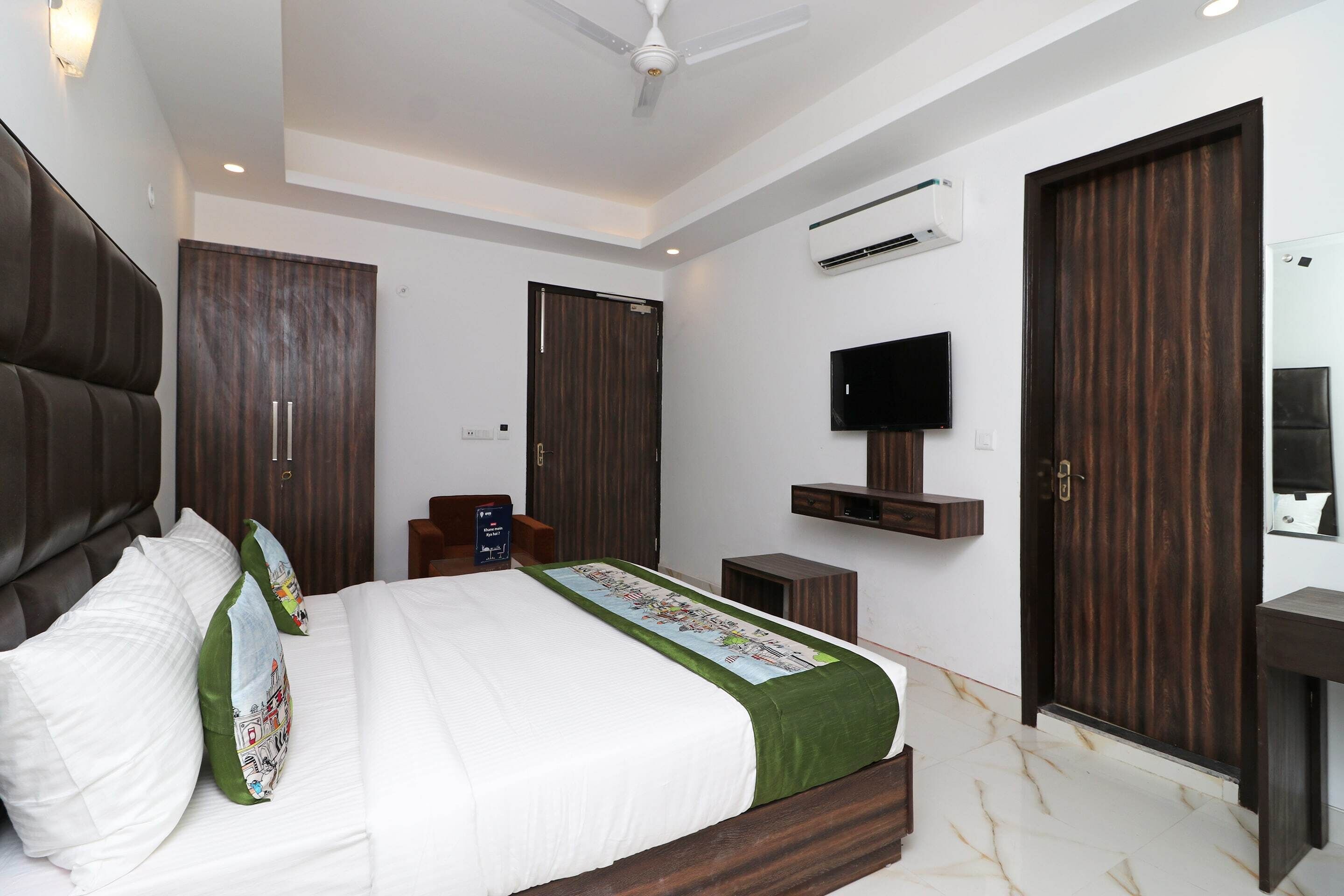 A modern bedroom with a large bed, dark wooden wardrobe, a mounted TV, and an air conditioning unit.