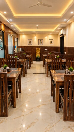 A restaurant with wooden tables and chairs, a central statue, and a ceiling fan. - Hotel Mayda Prime Near Delhi Airport (New Delhi)