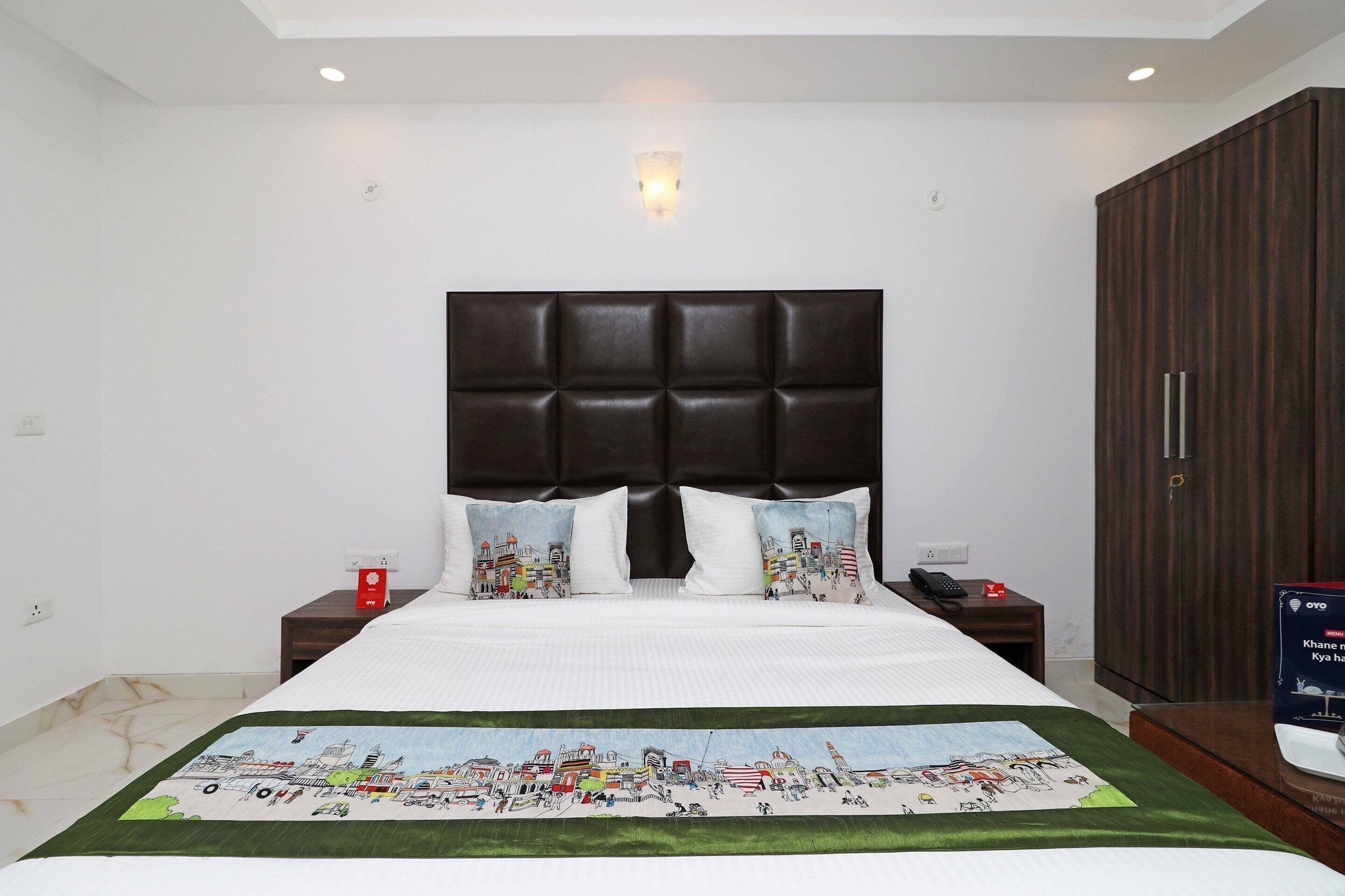 Una habitación de hotel moderna con una cama grande, un cabecero con diseño de paisaje urbano, una mesita de noche con lámpara y vistas de la ciudad desde la ventana.