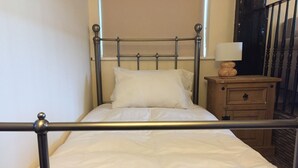 A single bed with a metal frame and white bedding, a wooden nightstand with a lamp, and a view of a balcony with a railing.
