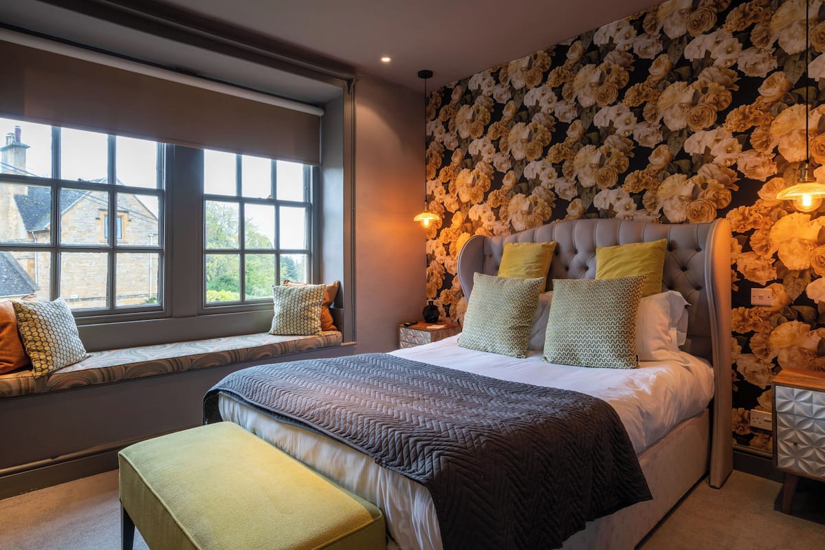 A bedroom with a large window, a bed with multiple pillows, and a floral wallpaper.
