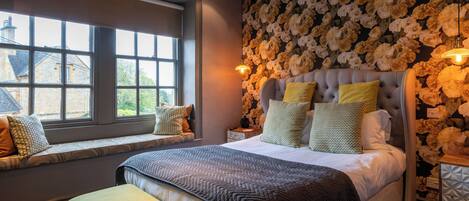 Une chambre avec une grande fenêtre, un lit doté de nombreux oreillers et un papier peint à motifs floraux.