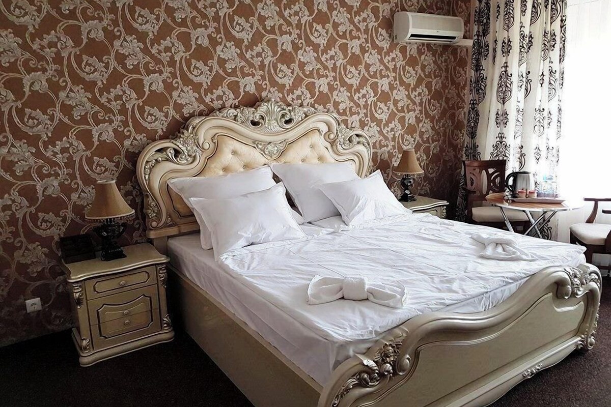 Une chambre d’hôtel avec un lit classique, une table de chevet, une lampe et une fenêtre avec des rideaux.