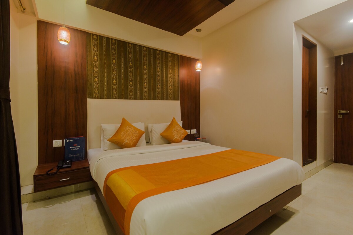 A neatly made bed with an orange blanket, wooden headboard, and two decorative pillows. A bedside table with a phone and a book is visible.