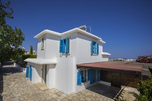 A white building with blue shutters and a red roof, featuring a stone-paved courtyard and a view of the sea. - Protaras Villa Ayios Elias Views (Paralimni)