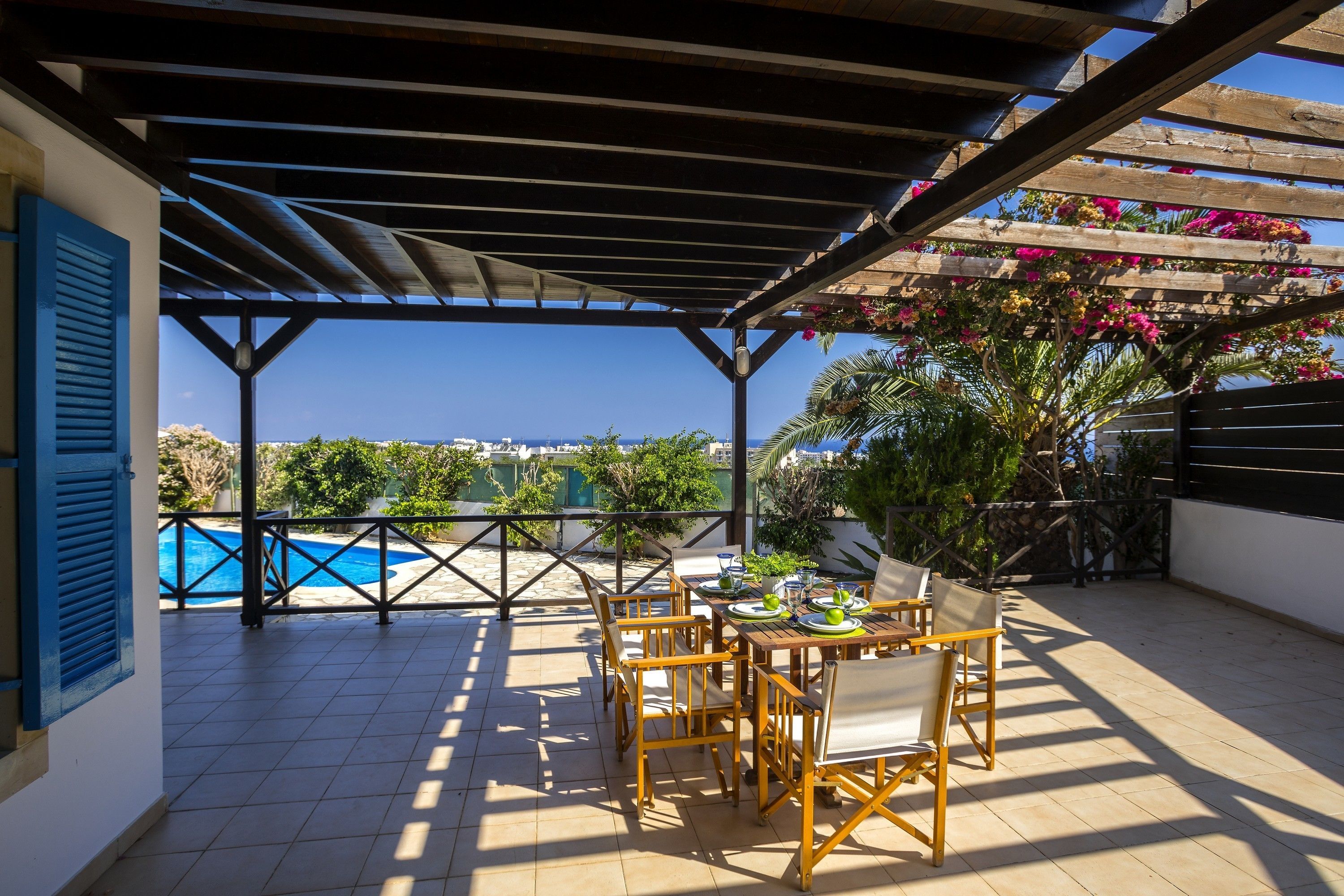 A covered patio with a pool view, wooden furniture, and a pergola with flowers.