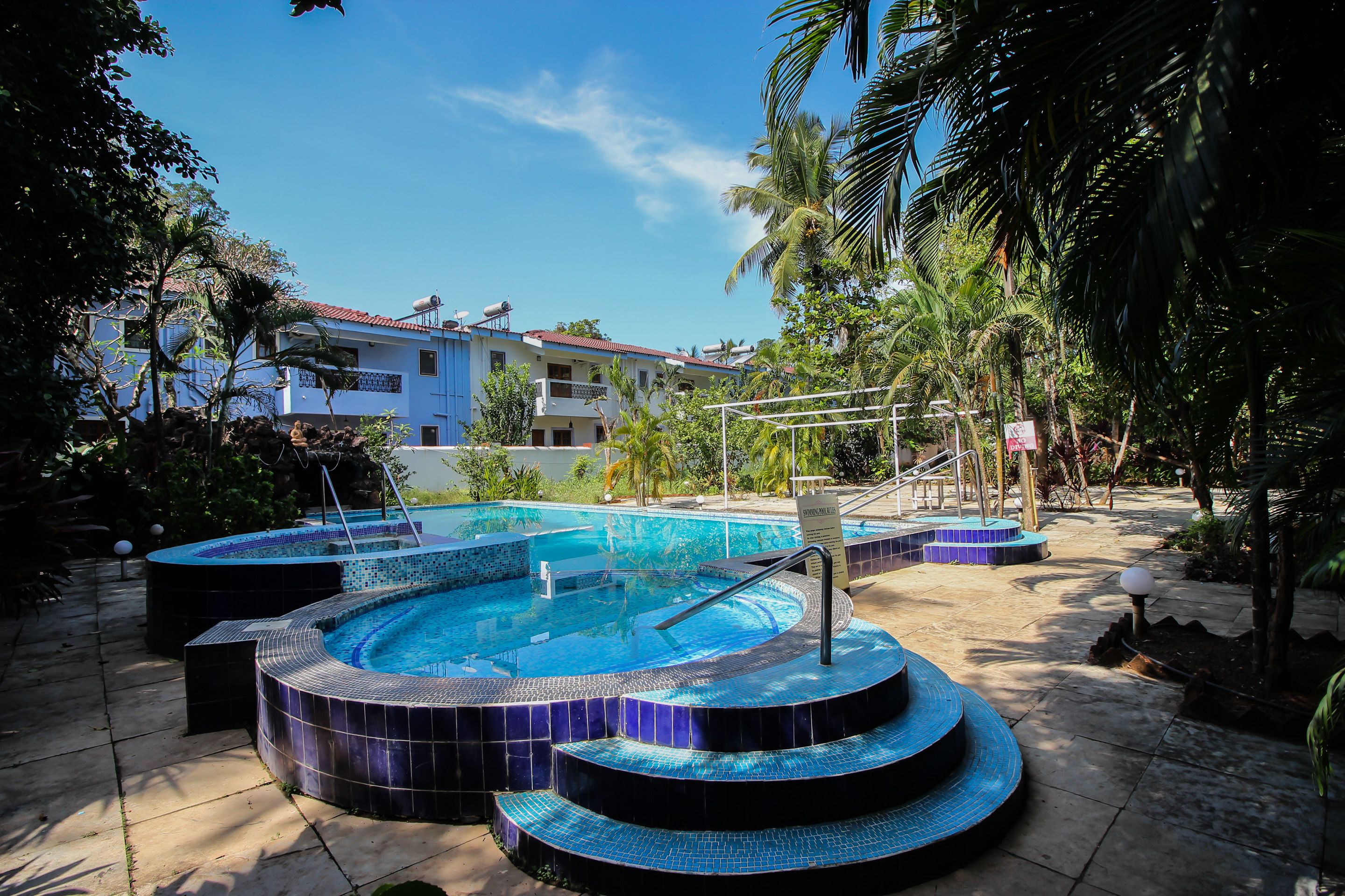 A swimming pool with a tiled edge and a small waterfall feature, surrounded by palm trees and a building with a red roof in the background.