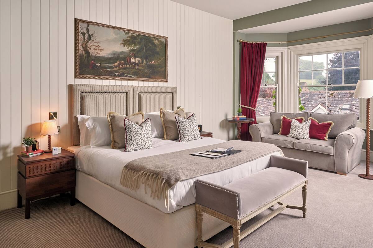 A bedroom with a large bed, a bedside table, a reading chair, a window with curtains, and a painting on the wall.