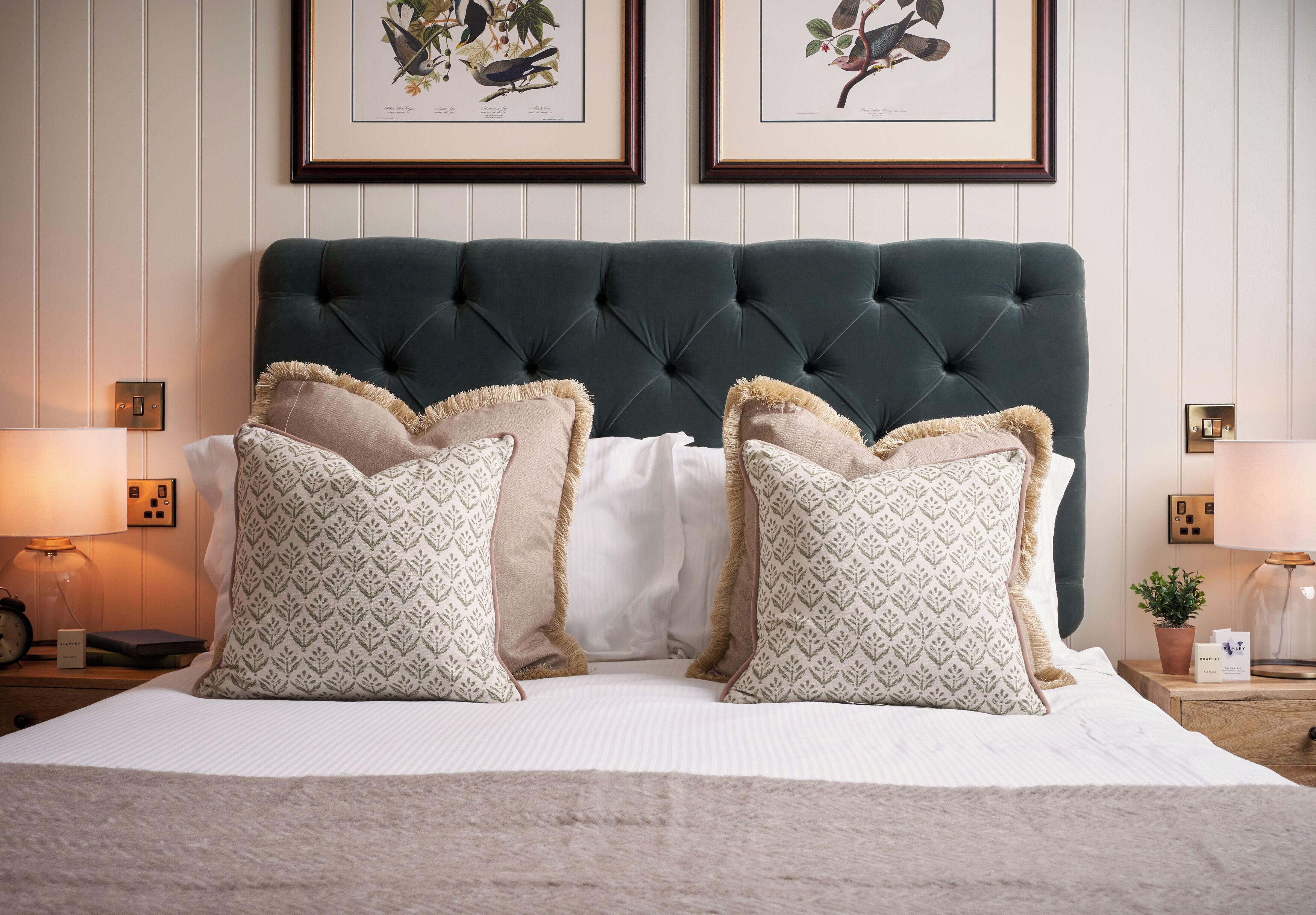 A neatly made bed with patterned pillows, a dark green headboard, and framed botanical prints on the wall.